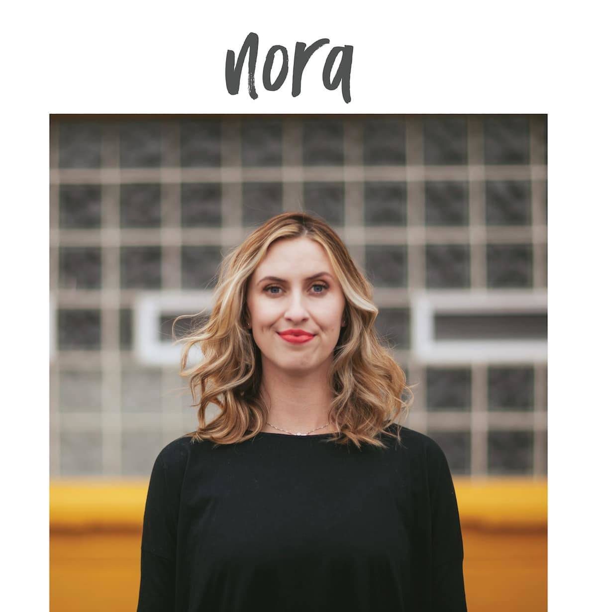A blond woman named Nora, standing with a half smile with a wearing a black top and red lipstick.